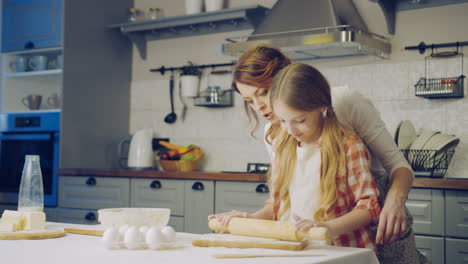 Beautiful-woman-and-her-little-daughter-making-a-daugh-for-the-cookies-on-the-kitchen-table-in-the-evening.-Indoors
